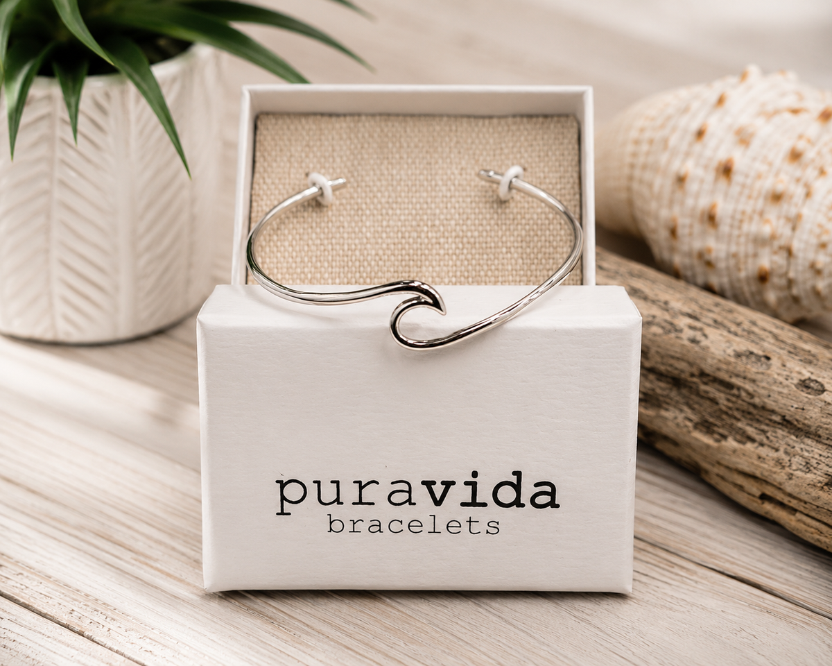 Pura Vida silver wave cuff bracelet displayed on its original gift box in a coastal-inspired setting with driftwood, seashells, and a decorative plant. The polished wave design is centered under soft natural lighting, highlighting the bracelet's sleek minimalist style.