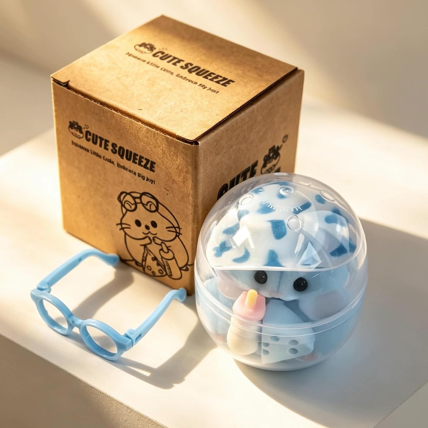Cute blue Taba squishy hamster wearing oversized blue glasses with rosy cheeks, accompanied by a miniature cheese wedge and baby bottle accessory, designed for stress relief, sensory play, and collecting.