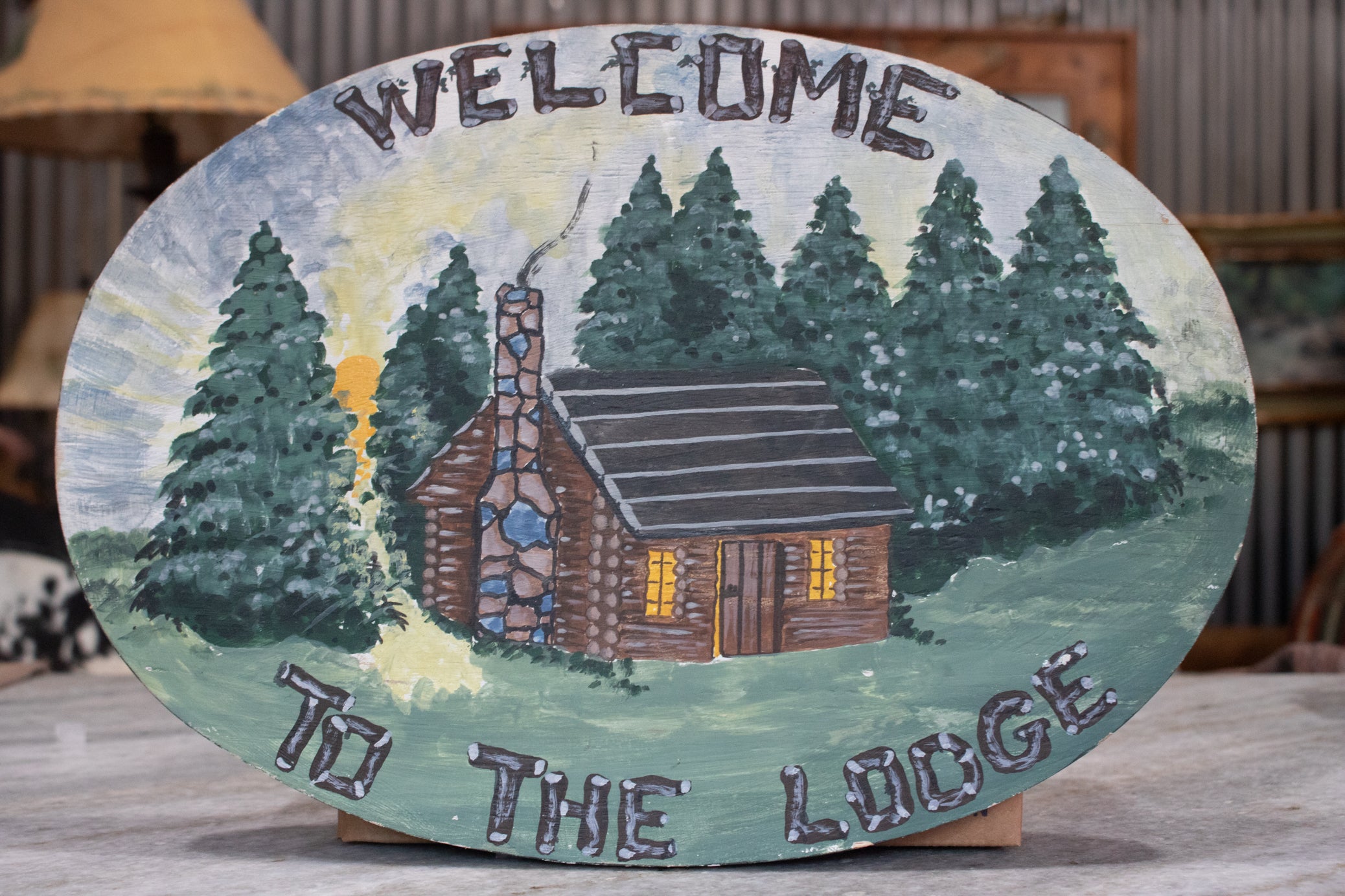 oval hand painted wooden welcome to the lodge wall sign featuring rustic cabin and pine tree forest scene with hanging hardware on back