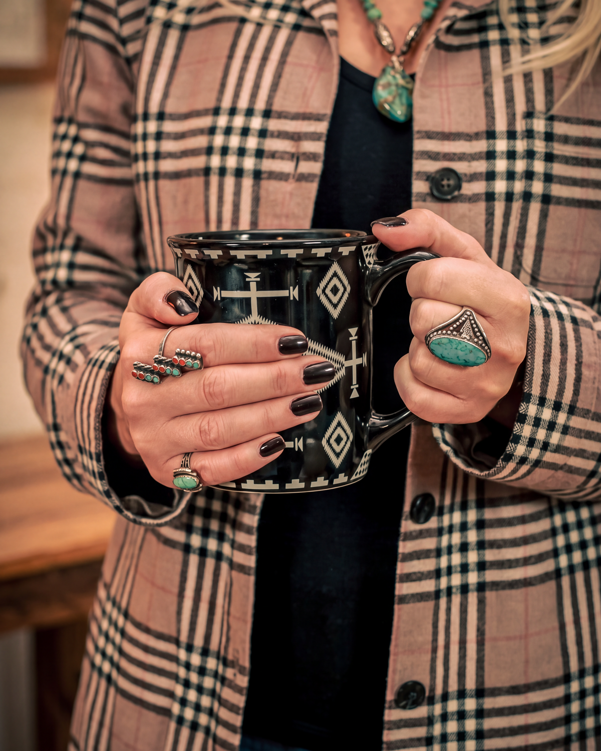 A person wearing a beige and black plaid jacket holds a black ceramic mug with a white geometric pattern. The person is also wearing turquoise jewelry, including a necklace and rings.